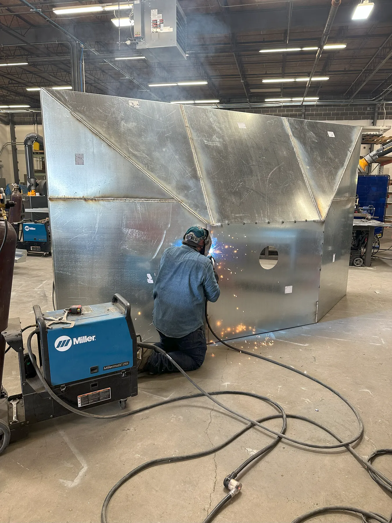 Expert Metal Fabrication and Welding for Custom Ductwork A welder using a Miller welding machine joins sections of large metal ductwork inside a fabrication shop, producing bright sparks during the process.